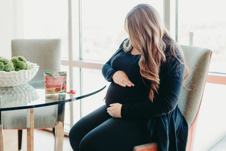 A pregnant woman sits at a glass table, gently touching her belly with her right hand. A package of morning wellness tea and a red tea bag, some of the best products for a better pregnancy, rest on the table. The background features a bowl of green decorative items and large windows with natural light.