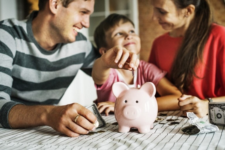 A family is gathered around a table, smiling. The child is placing coins into a pink piggy bank while the adults, presumably the parents, look on. There are dollar bills and coins scattered on the table, perhaps part of their monthly family budget planning.