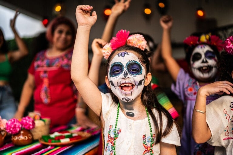 A group of children and adults, some with painted faces depicting sugar skull designs, celebrate. The scene includes bright decorations, including flowers and textiles. One child in front, with face paint and flowers in her hair, has her arm raised and is smiling broadly next to a colorful shoebox altar.