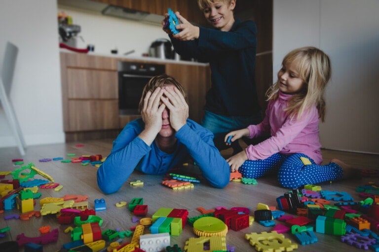 An adult lies on the floor covering their face, likely feeling burnout, surrounded by scattered colorful building blocks. Two children, one standing with a block above the adult's head and the other sitting next to the adult, are engaged in play in a kitchen.