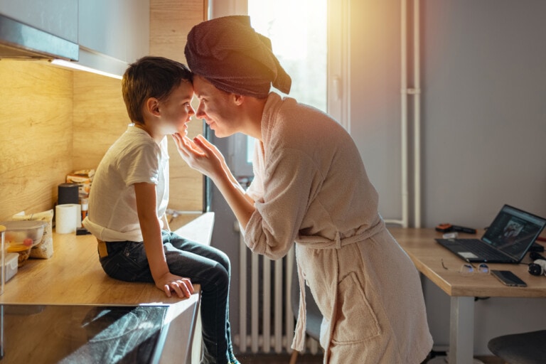 An adult wearing a bathrobe and a towel wrapped around their head is gently touching noses with a child sitting on a kitchen counter, perhaps sharing moments of positive self-talk for kids. The background shows a kitchen with sunlight streaming through a window and a computer on the desk.