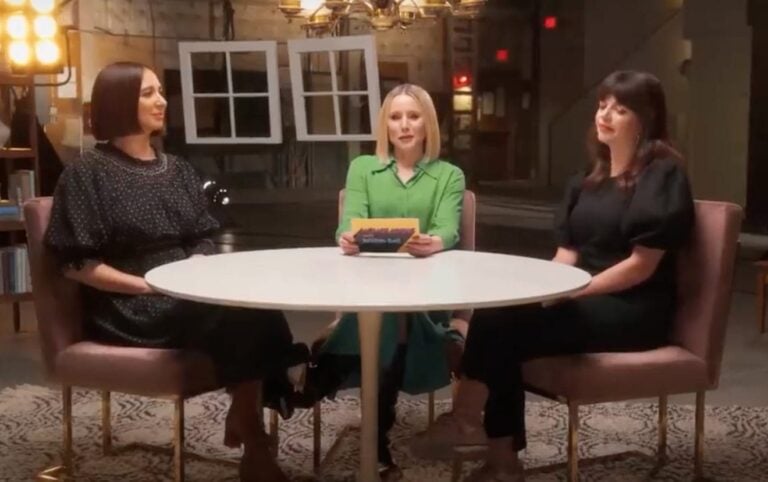 Three women, including Kristen Bell, are seated around a circular table in a well-lit room. The woman in the center is holding a card, while the other two listen attentively. They are all dressed in dark or neutral-toned clothing. The background features industrial-style decor.