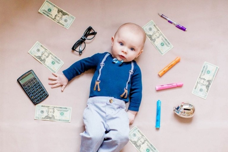 A baby dressed in a blue outfit with suspenders lies on a pink surface surrounded by dollar bills, a calculator, glasses, a pen, markers, and a small globe. With a neutral expression facing the camera, this setup hints at tips to save money on baby costs.