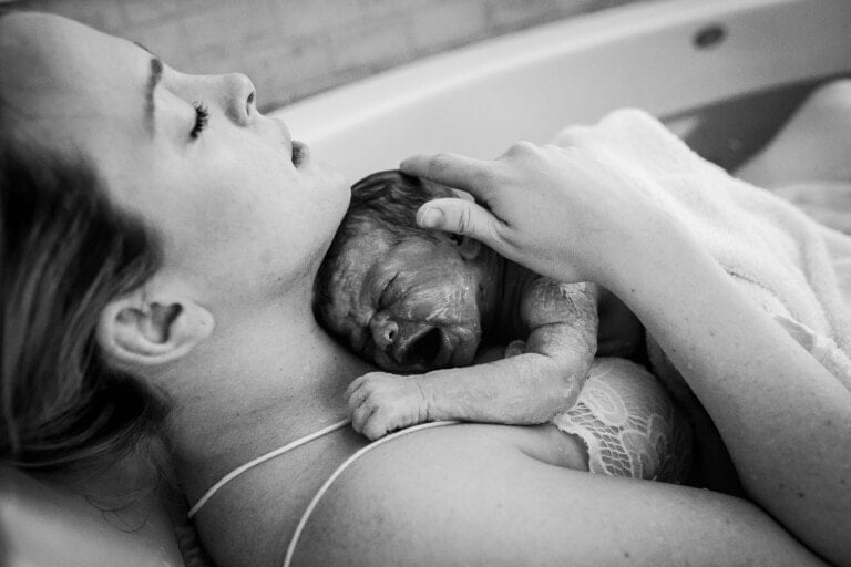 A woman in a bathtub holds a newborn baby against her chest after a vaginal birth. The baby is crying, and the woman's eyes are closed as she rests her head back. A towel partially covers the newborn. The scene appears to be a home water birth.
