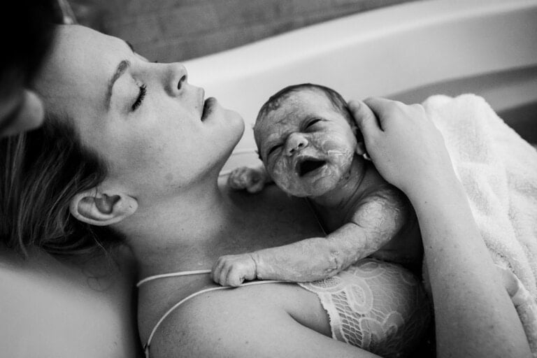 A woman in a white lace top cradles a newborn baby in her arms while sitting in a bath, experiencing the aftermath of our home birth. The baby, still covered in vernix, has its eyes open and mouth slightly open. The woman appears to be resting with her eyes closed.