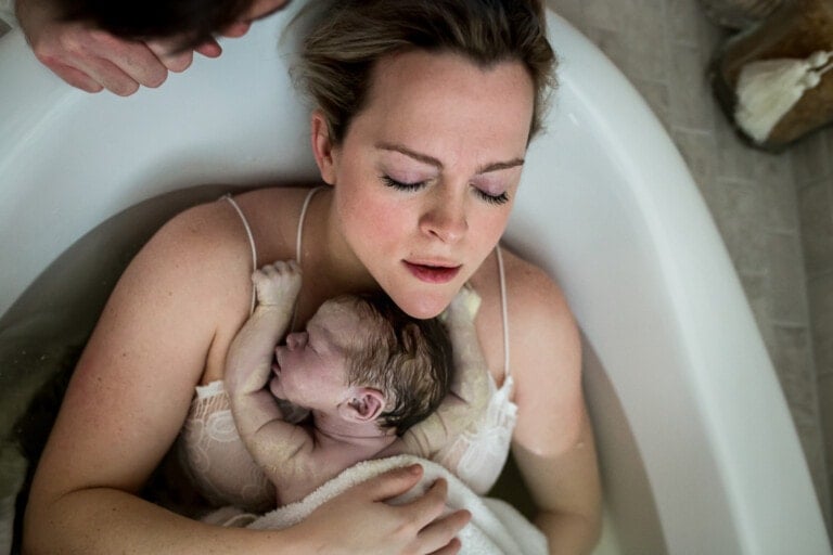 A woman, eyes closed, is lying in a birthing tub holding a newborn baby against her chest, embracing the beauty of a water birth. The baby appears to be freshly born, with damp hair and vernix on its skin. A person, partially visible, is leaning over the tub.