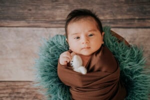 A baby wrapped in a brown blanket lies on a furry green cushion. The baby is holding a small white plush bear and looking up at the camera, as if contemplating s names for boys. The background features wooden planks, adding a rustic feel to the scene.