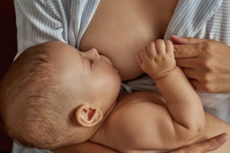 A baby is breastfeeding, held closely by a person wearing a light blue and white striped shirt. The baby's eyes are closed and one hand rests gently against the person's chest, embodying the warmth and connection celebrated in breastfeeding affirmations.