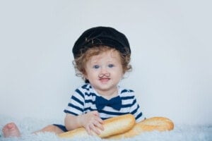 A toddler with curly hair is wearing a striped shirt, black beret, and blue bow tie. They have a small painted-on mustache and are holding two baguettes, sitting on a soft white surface with a neutral background. This little one could easily be named Louis or Camille from classic French boy names.