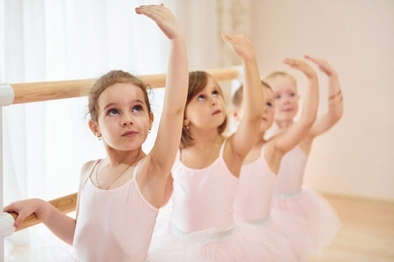 Four young girls in pink ballet outfits stand in a line at a ballet barre, gracefully raising one arm as they practice their dance activities. They are in a brightly lit dance studio.