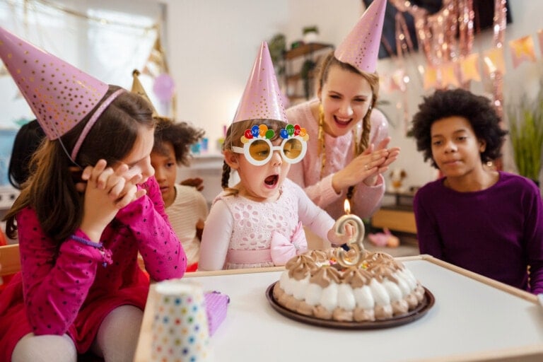 A young child wearing colorful glasses and a birthday hat gasps while looking at a cake with a lit number 3 candle. Two children and an adult wearing party hats surround the child, clapping and smiling in a decorated room, ensuring all treats are safe for food allergies.