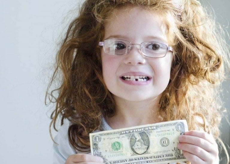 A young child with curly hair and glasses smiles, showing a gap where a tooth is missing—clearly thrilled with the successful implementation of one of those imaginative tooth fairy ideas. The child is holding a one-dollar bill in both hands, set against a plain and neutral background.