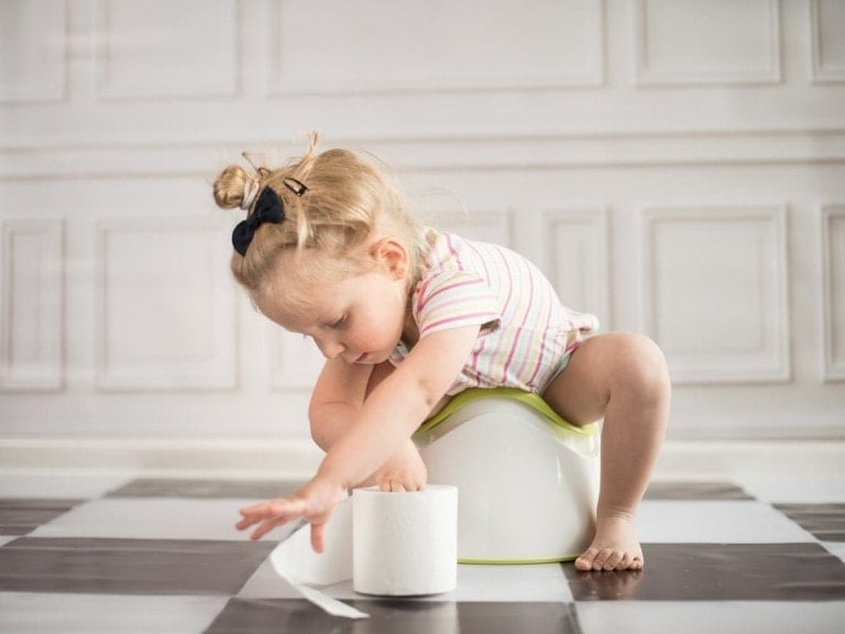 A young child with blonde hair sits on a potty training seat, reaching for a roll of toilet paper on the tiled floor. The child is wearing a striped short-sleeved shirt. The background features white, paneled walls—a perfect scene featuring essential potty training must haves.