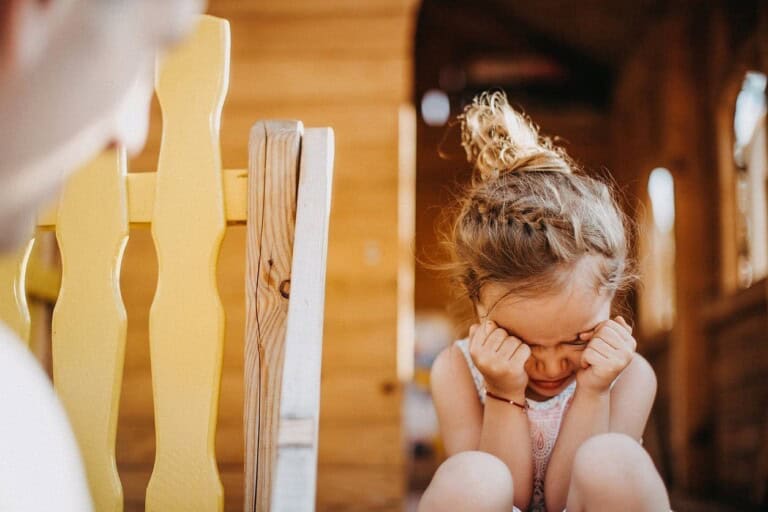 A young child with braided hair sits by a yellow wooden rail in the sunlight, covering their eyes with their hands, seemingly upset. Its a tender moment that reminds us of the importance to nurture understanding and patience as you unspoil your child.