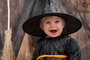 A baby dressed in a black witch costume with an orange belt smiles widely. The baby is wearing a large pointed witch hat. There is a straw broomstick on the left side of the image and a wooden background draped with dark fabric, adding to the enchantment of this scene perfect for any witchy names.