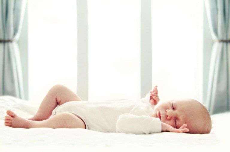 A baby in a white onesie sleeps on its back on a white bed. The baby's right arm is up by its head, and its left arm is resting on its torso. Light blue curtains and a bright window are in the background. When can a baby sleep on their stomach safely? Usually, after they can roll over consistently by themselves.