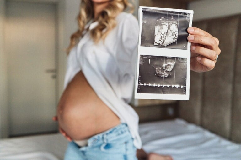 A pregnant woman is standing in a room, holding up an ultrasound image in her right hand. She is wearing a white blouse and unbuttoned jeans. The ultrasound shows two separate images, revealing she is pregnant with twins. The woman’s face is not visible in the photo.