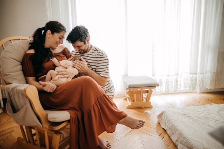 A woman in a long dress sits in a rocking chair holding a baby, while a man kneels beside them, smiling and touching the baby's head. They are in a sunlit room with wooden flooring and sheer curtains, having refused visitors to cherish this intimate moment. Another chair and a low bed are visible in the background.
