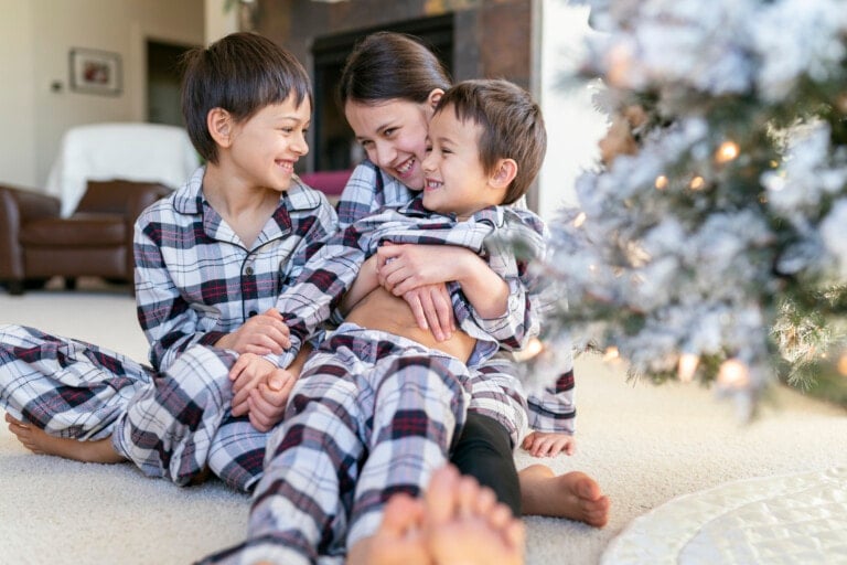 Three children wearing matching family holiday pajamas sit on the floor near a Christmas tree. They are smiling and laughing, with one child sitting in the middle and hugging the other two. The scene appears to be festive and playful.