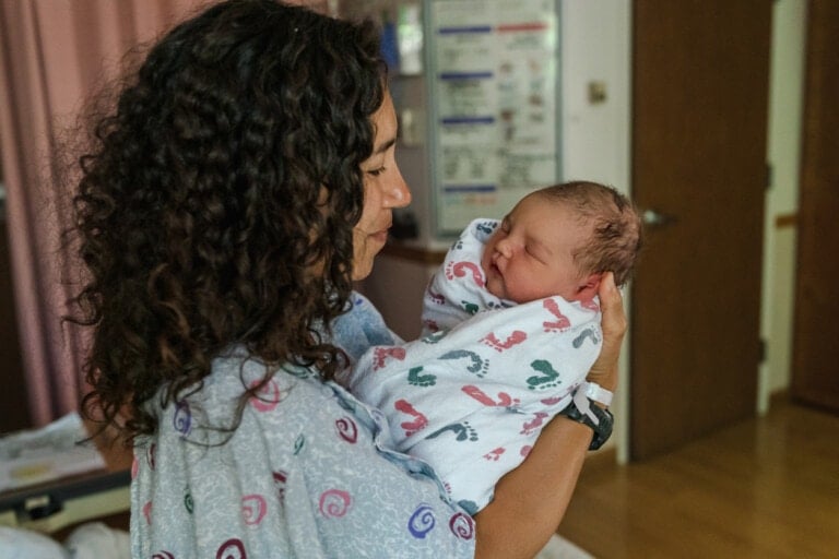 A woman with curly hair holds a newborn wrapped in a white blanket adorned with colorful hand and foot prints. They are in a room with a brown door and medical posters detailing birthing positions visible in the background.