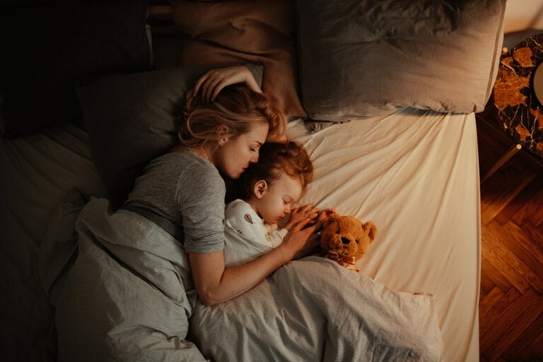 A woman and a child lay peacefully asleep in bed, exemplifying co-sleeping parents. They rest close to each other under a gray blanket, the child clinging to a brown teddy bear. The bed is adorned with gray and beige pillows, while a bedside table with a lamp adds a soft glow in the background.