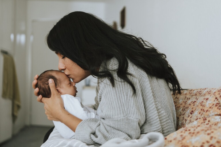 A woman with long dark hair holds and kisses the forehead of a baby, finding solace and pain relief after childbirth. She is seated on a patterned sofa in a neutral-toned room. The baby is dressed in white, and both appear affectionate and calm.