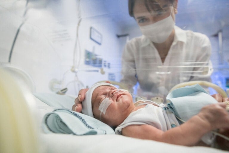 A NICU baby lies in an incubator with medical tubes attached while an adult wearing a white mask and shirt looks through the glass, placing a hand on the baby's head. The scene appears to take place in a neonatal intensive care unit.