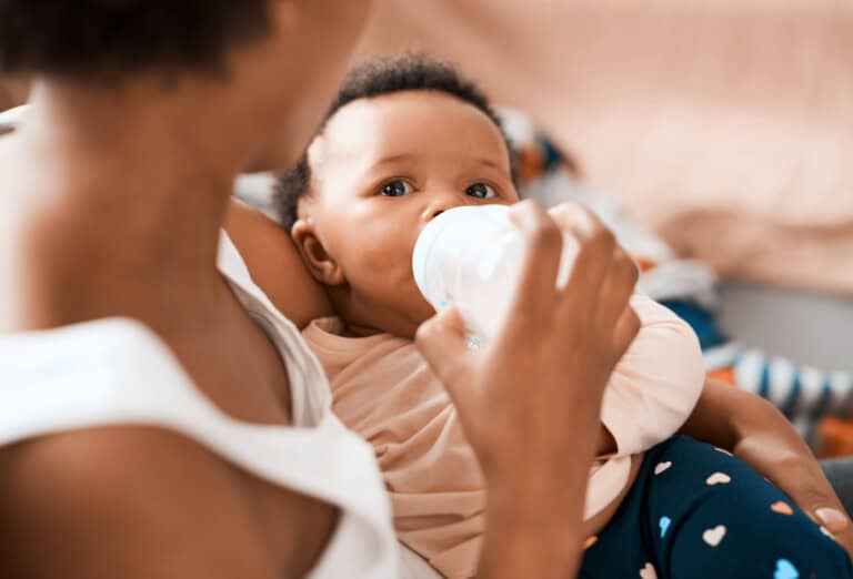 A mother holds her baby who is drinking from a bottle, wearing a light pink shirt and dark blue pants with white spots. The woman's face is not visible as she looks down, attentive to any baby full cues. The background is blurry with warm-toned furnishings.