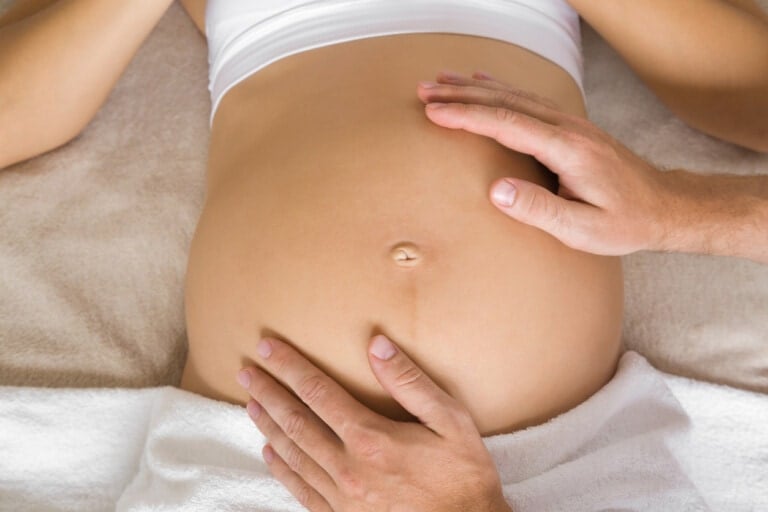 A close-up image of a pregnant woman laying on her back with her midsection exposed. Two hands, likely belonging to another person, are placed gently on her belly, one above the belly button and one below, perhaps feeling for a breech baby. The woman is covered with a white towel from the waist down.