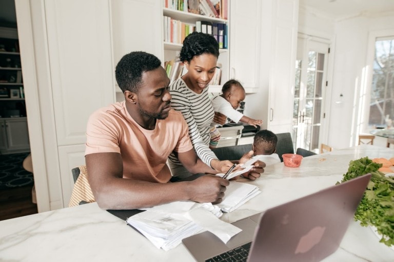 A man and woman are in a kitchen, with the man looking at papers and the woman holding a baby. Another child is sitting at the table with toys. A laptop is open on the countertop, surrounded by vegetables. Books on growing your money are visible on a shelf in the background.