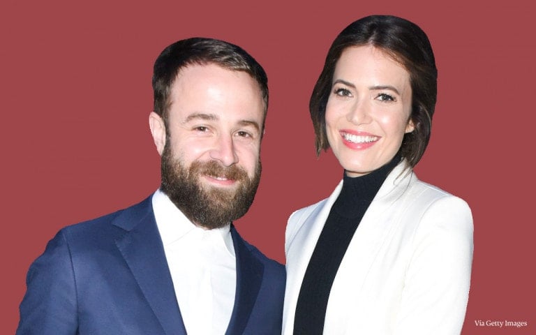 A man and woman are posing together in front of a solid maroon background. The man has a beard and short hair, wearing a navy blue suit with a white shirt. The woman, with shoulder-length dark hair, dons a white blazer over a black top. "Via Getty Images" text is in the bottom right corner, evoking the elegance often seen in Mandy Moore's photoshoots.