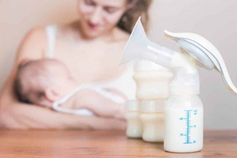 A person holding an infant is visible in the background, while a manual breast pump and several filled milk bottles are placed on a wooden surface in the foreground. The image focuses on the breast pump and milk bottles, highlighting how to store breast milk for future use.