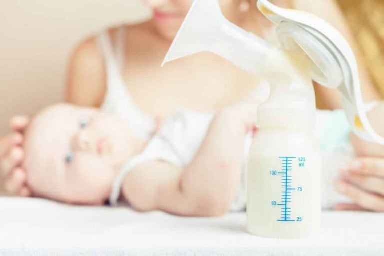 A baby lies in the background with an adult holding them. In the foreground, a breast pump with a bottle of expressed milk, showcasing measurement markings, suggests tips on how to increase milk supply. The scene appears to be set in a domestic environment.