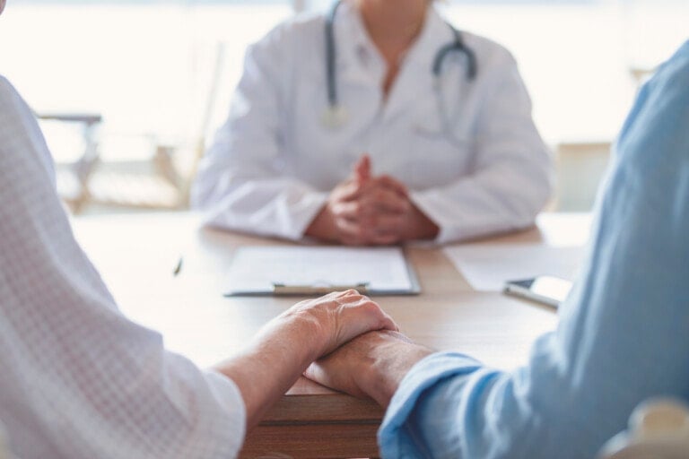 An older couple holds hands at a table while a doctor, dressed in a white coat with a stethoscope, sits across from them with a clipboard and a tablet. The setting appears to be a consultation or medical discussion about fertility treatments in a bright room.