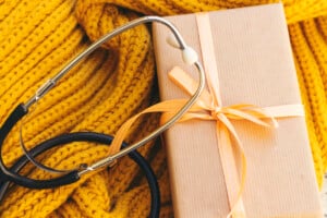 A stethoscope rests on a yellow knitted fabric beside a brown paper-wrapped gift box tied with a yellow ribbon. The objects are arranged closely, suggesting a theme that might be related to gifts for labor delivery nurses or celebrating the dedication of medical professionals.