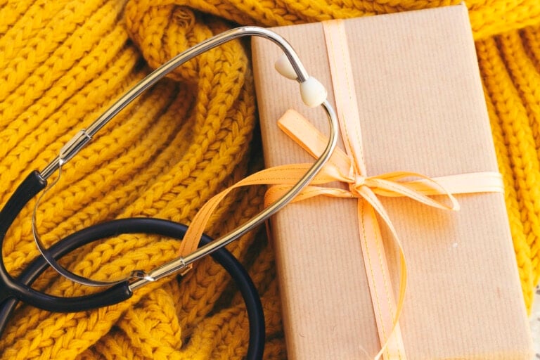 A stethoscope rests on a yellow knitted fabric beside a brown paper-wrapped gift box tied with a yellow ribbon. The objects are arranged closely, suggesting a theme that might be related to gifts for labor delivery nurses or celebrating the dedication of medical professionals.
