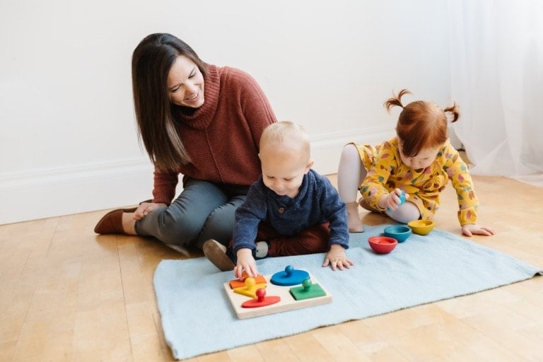An adult sitting on the floor watches two young children playing. One child is working on a shape sorter puzzle, and the other is playing with colorful stacking toys. The scene, reminiscent of a Montessori podcast, is set in a brightly lit room with hardwood floors.