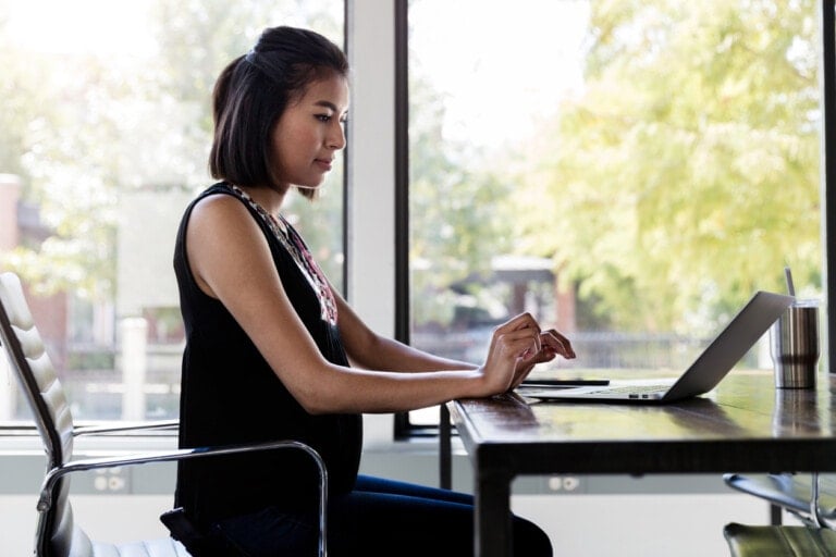A person with shoulder-length hair, working during pregnancy, sits at a desk and types on a laptop. They are in a bright room with large windows that show trees and sunlight outside. An insulated tumbler sits next to them on the wooden desk.