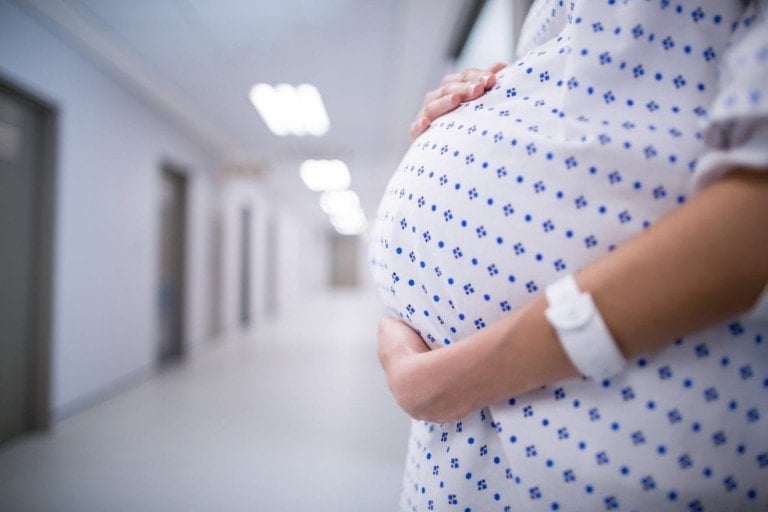 A pregnant person wearing a hospital gown and wristband stands in a well-lit, sterile hospital corridor, holding their belly. Amid the covid pandemic, the sense of anticipation is palpable as they prepare to have their baby in this clean environment with closed doors lining the walls.