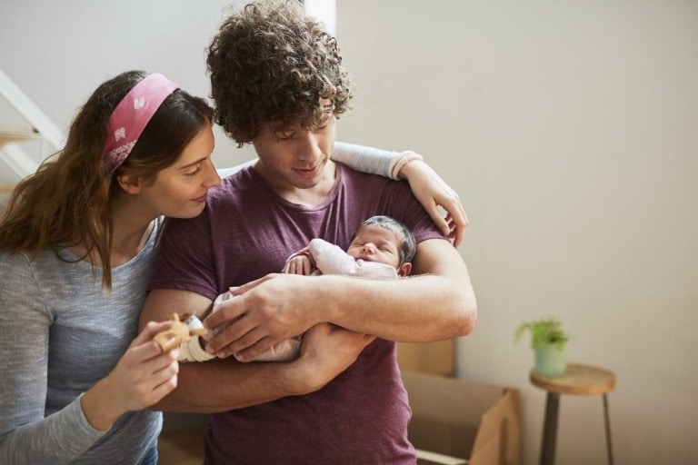 A man holds a newborn baby in his arms while a woman stands close, looking at the baby. The woman has her arm around the man's shoulder and holds a toy giraffe. They are indoors, possibly in a living room with a potted plant in the background, reflecting their practical approach to staying on budget.