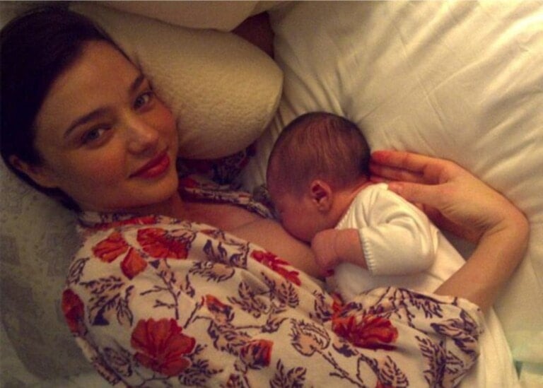 A woman is lying down in bed, breastfeeding an infant. She is wearing a floral-patterned robe and smiling at the camera like one of those radiant celebrity moms. The baby is dressed in a white outfit.