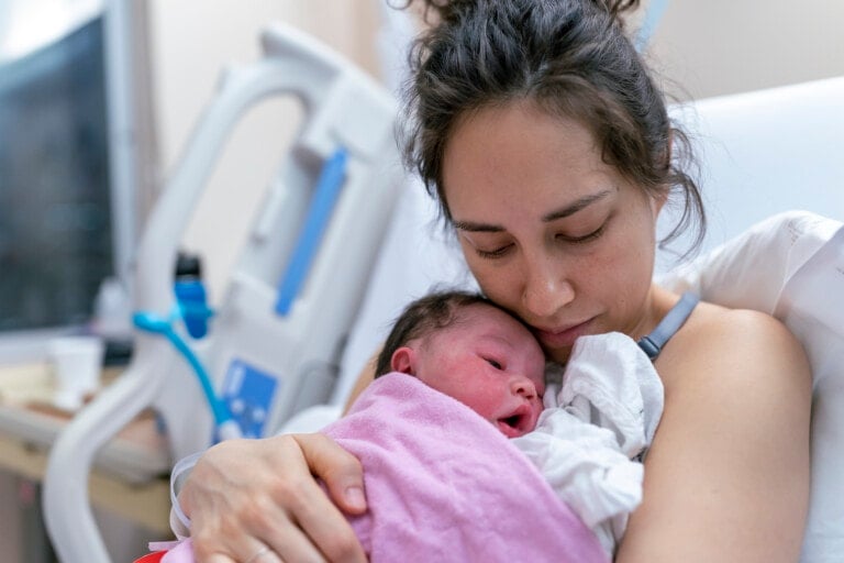 A woman with dark hair tied up is in a hospital bed, holding a newborn baby wrapped in a pink blanket against her chest. Despite the need to mourn your birth plan, she looks down lovingly at the baby. Medical equipment is visible in the background.