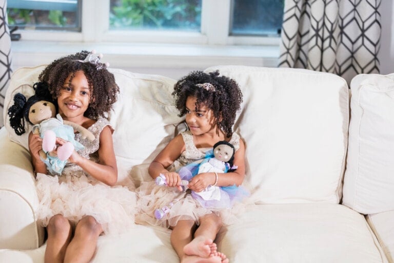 Two young girls sit on a white couch by a window, each holding diverse dolls. Both are wearing matching beige dresses and have curly hair. One girl smiles at the camera, while the other looks down at her doll. Patterned curtains provide a charming backdrop.