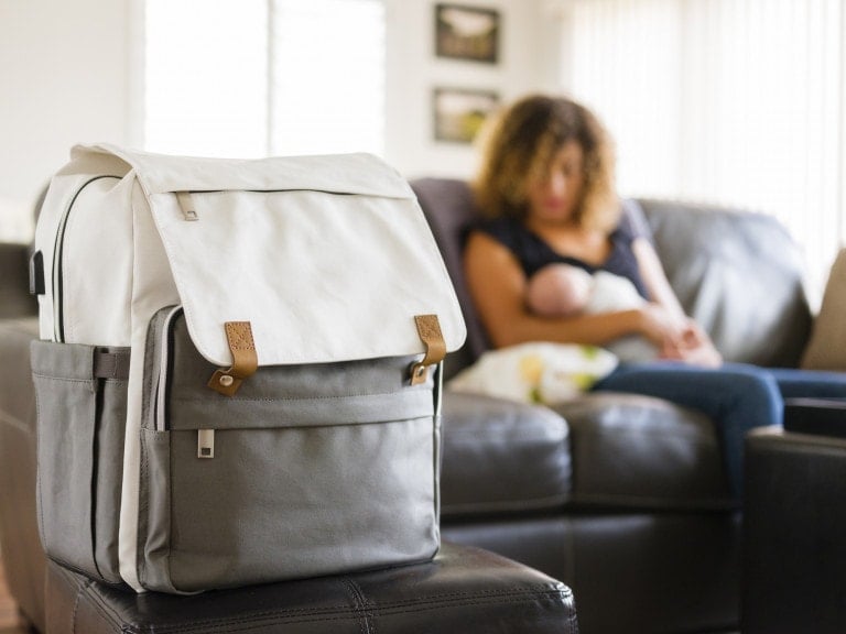 A white and gray diaper bag with brown strap accents is placed on a black ottoman in the foreground. In the background, a woman with curly hair is sitting on a couch, breastfeeding a baby surrounded by toys. The room is brightly lit with a window and picture frames on the wall.