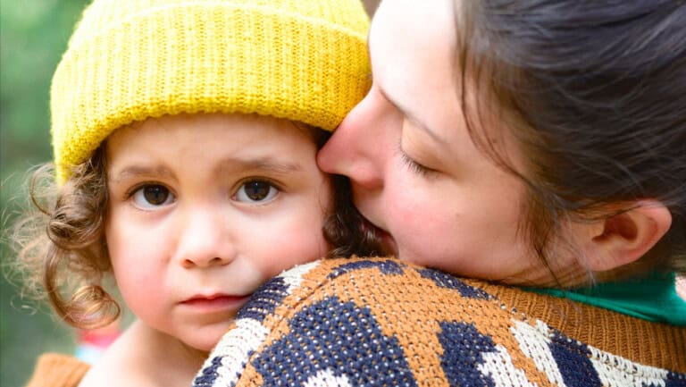 A young child with curly hair, possibly in the throes of the terrible twos, wears a yellow knit hat and is held closely by an adult wrapped in a patterned sweater. The adults eyes are closed, cheeks gently touching the childs. The background is blurred and green.