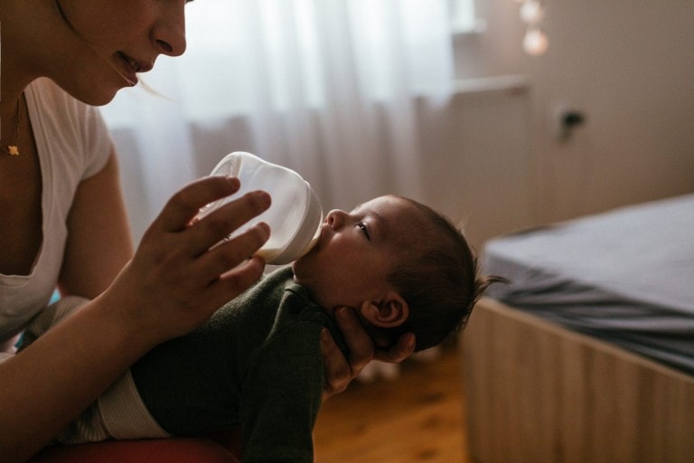 A person is trying to feed an infant with a bottle in a dimly lit room, but the baby won't take the bottle. The infant, wearing a green outfit, lies in the person's arms. A bed and white curtains are visible in the background.