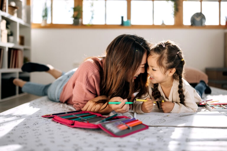 A woman and a young girl lie on their stomachs on a bed, smiling and looking at each other while holding colored pens. The bed has a white bedspread with black patterns, and a set of colored pens is open in front of them along with some paper. It's the perfect scene that captures the essence of parenting.