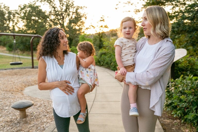 Two women are walking with their young children along a path in a park, discussing playdate questions. One woman is pregnant and carrying a toddler, while the other is holding a child. Trees and a swing set are visible in the background.