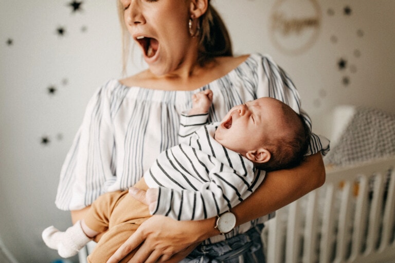 A person, possibly trying to help the tired mom, is holding a yawning baby while yawning themselves. The person is wearing a striped shirt and hoop earrings, and the baby is dressed in a striped onesie with tan pants. Background includes wall decorations and a crib.