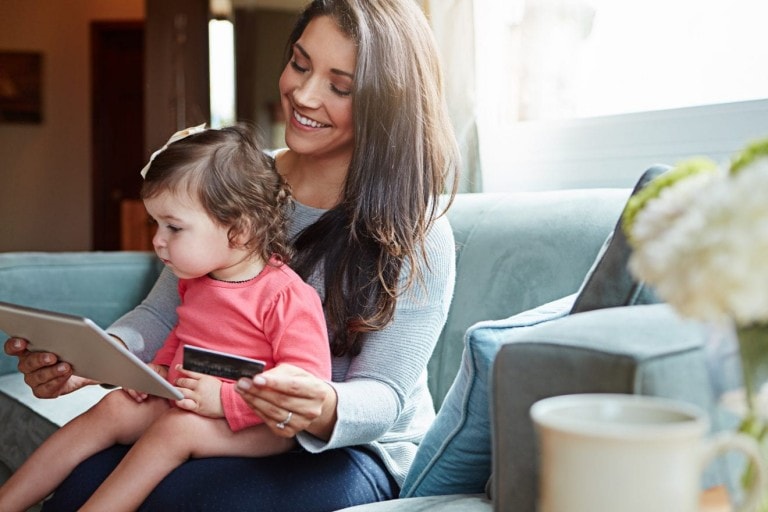 A woman sits on a couch holding a tablet and a credit card, while a small child sits on her lap intently looking at the tablet screen. The scene is indoors with soft focus background that includes a window and some flowers, illustrating when to use a credit card for easy online shopping.